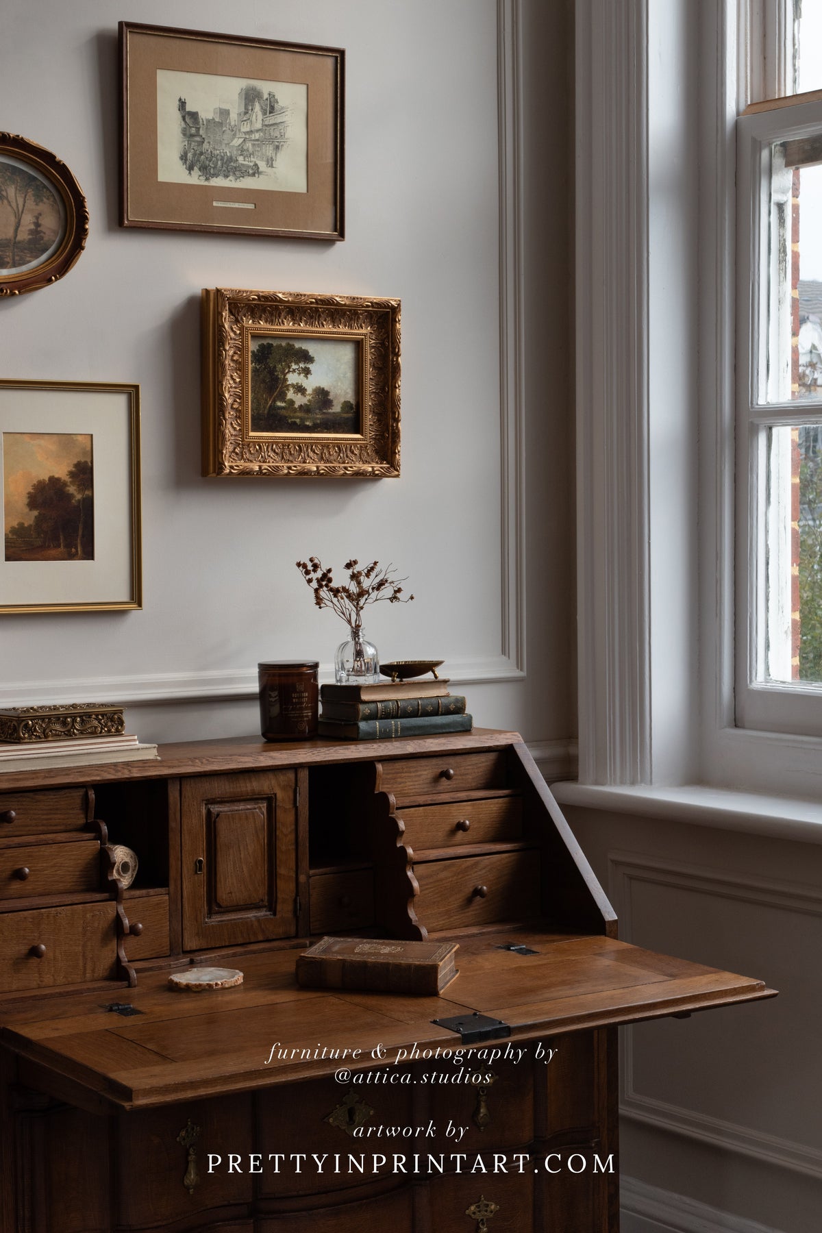 Antique oak bureau beneath a small gallery wall of vintage landscapes in gold and wood frames, beside a tall sash window