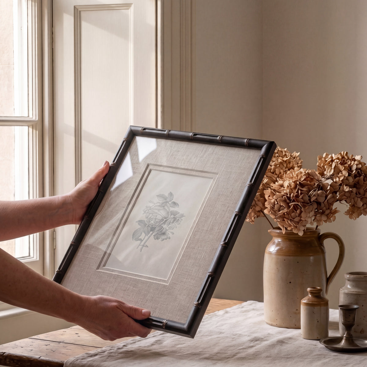 Hands holding a framed vintage wall art print above a table styled with decorative antique pieces