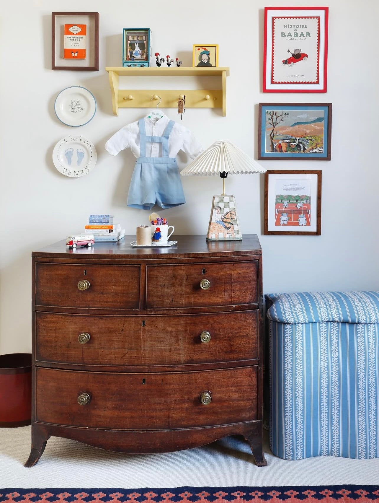Nursery corner with a vintage dresser, children's books, toys, and wall art including our framed Babar nursery print in red. A romper hangs below a yellow shelf with figurines, and a blue striped ottoman sits nearby.
