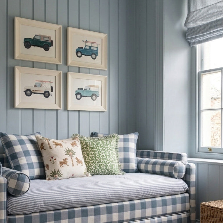 A blue and white gingham upholstered daybed in a styled room with blue paneled walls, four framed car prints, patterned pillows, a rug, and toys near a window.