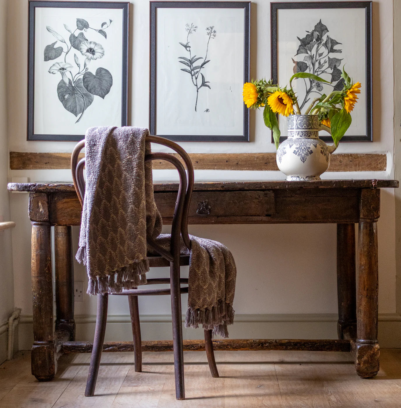 Rustic antique wooden writing desk and bentwood chair styled with vintage botanical art prints, a textured throw, and sunflowers in a patterned ceramic pitcher.
