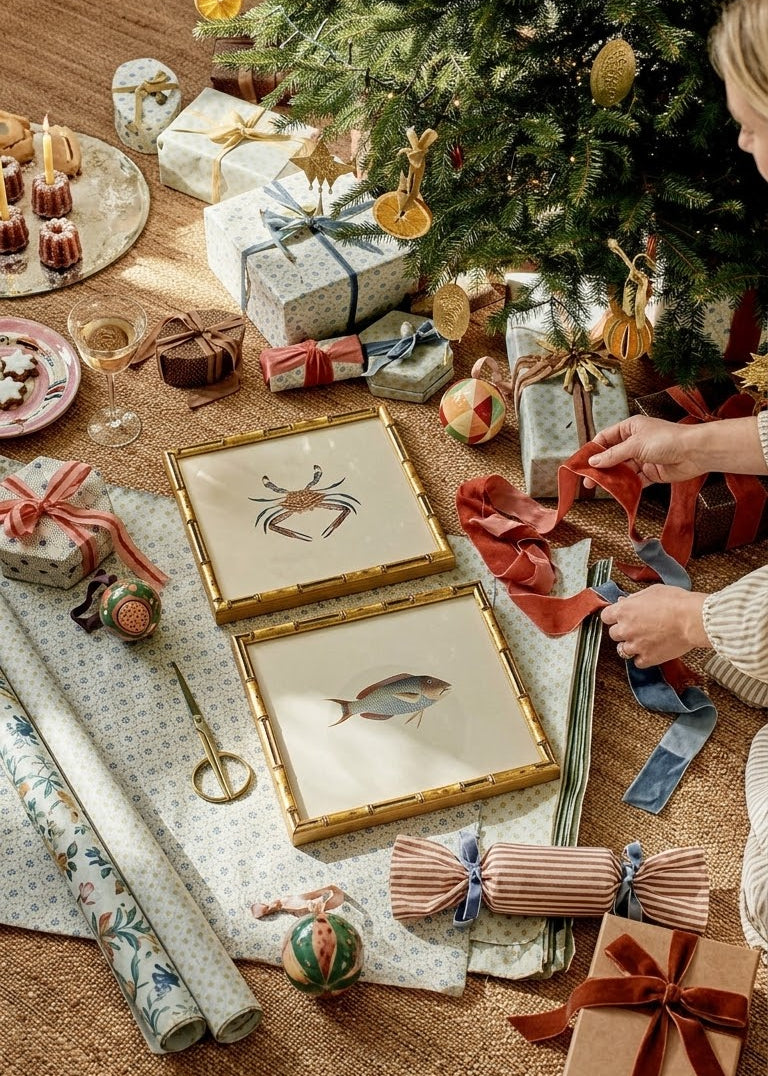 A cozy holiday scene with a decorated Christmas tree, wrapped gifts, and two framed art prints featuring a crab and a fish on a table. A person's hands and arm are visible wrapping a gift, creating a warm, festive atmosphere.