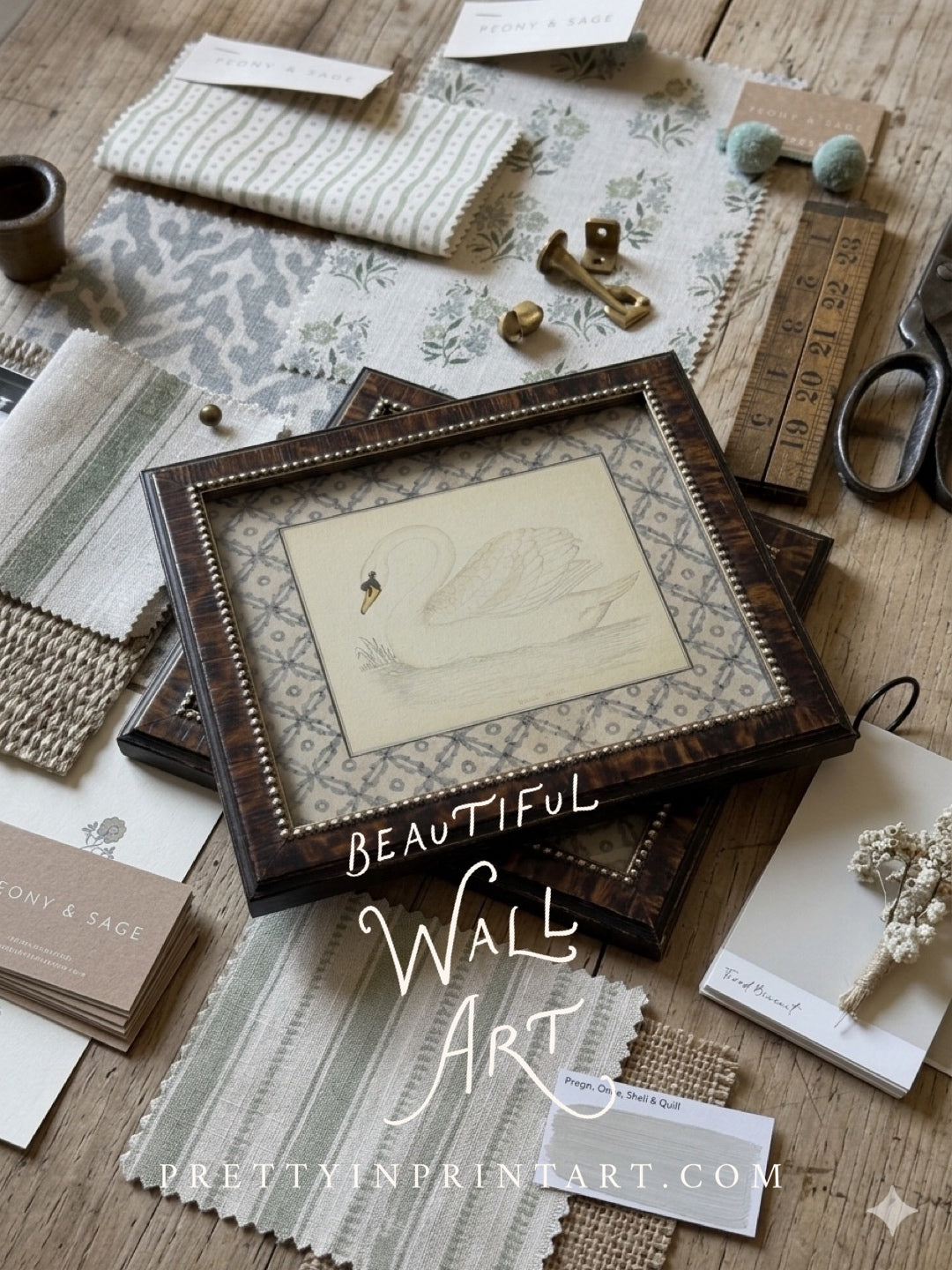 A styled flat-lay of framed vintage swan wall art prints and samples of timeless fabrics on a wooden surface. The central product appears to be a square framed print or artwork sample with a dark wood frame, cream background, and a delicate swan illustration.