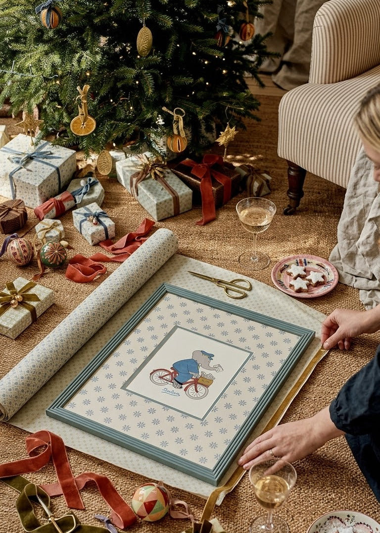 framed babar nursery art, showing babar the elephant on his bike dressed as a postman. In blue frame painted in Oval Room Blue by Farrow & Ball. Christmas scene at home, surrounded by wrapping paper, velvet ribbons, idyllic Christmas home Christmas 