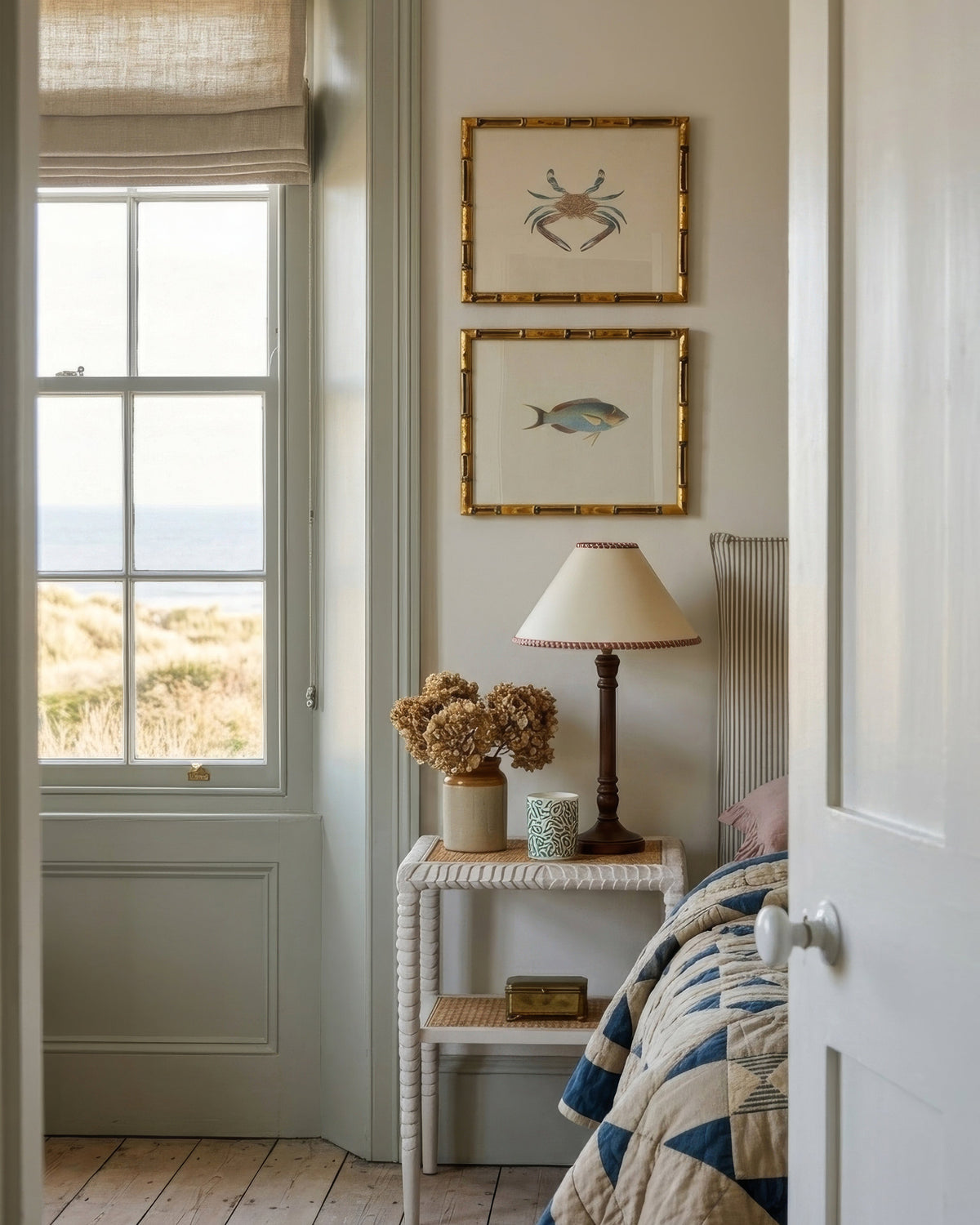 A styled bedroom interior with a window, framed crab and fish artwork on the wall, a bedside table with a lamp and flowers, and part of a quilted bedspread visible in a softly lit room.