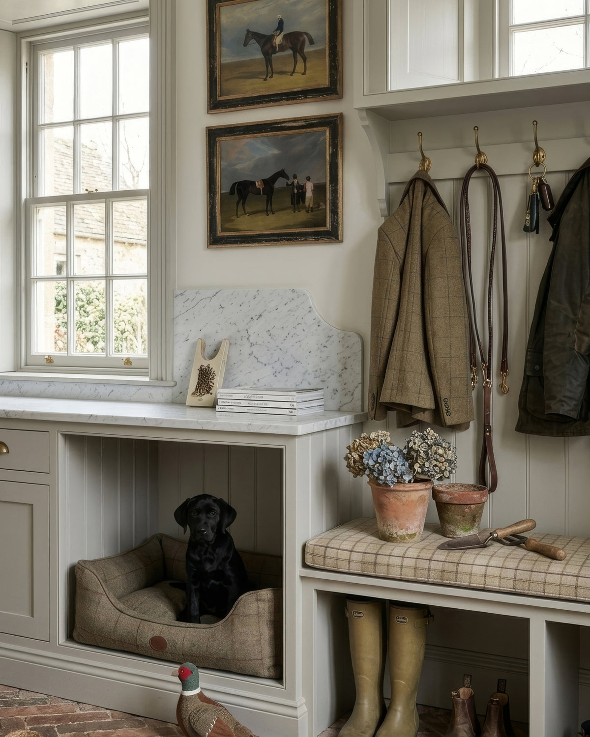 A rustic country-style mudroom featuring a built-in bench with a plaid cushion, green rain boots, horse paintings on the wall, and a dog sitting in a pet bed under a window.