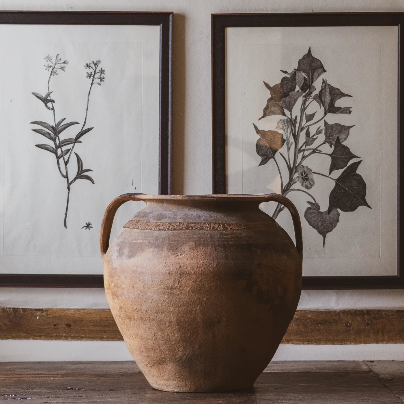 Vintage Brown ceramic vase on a antique wooden surface with framed botanical prints in the background.